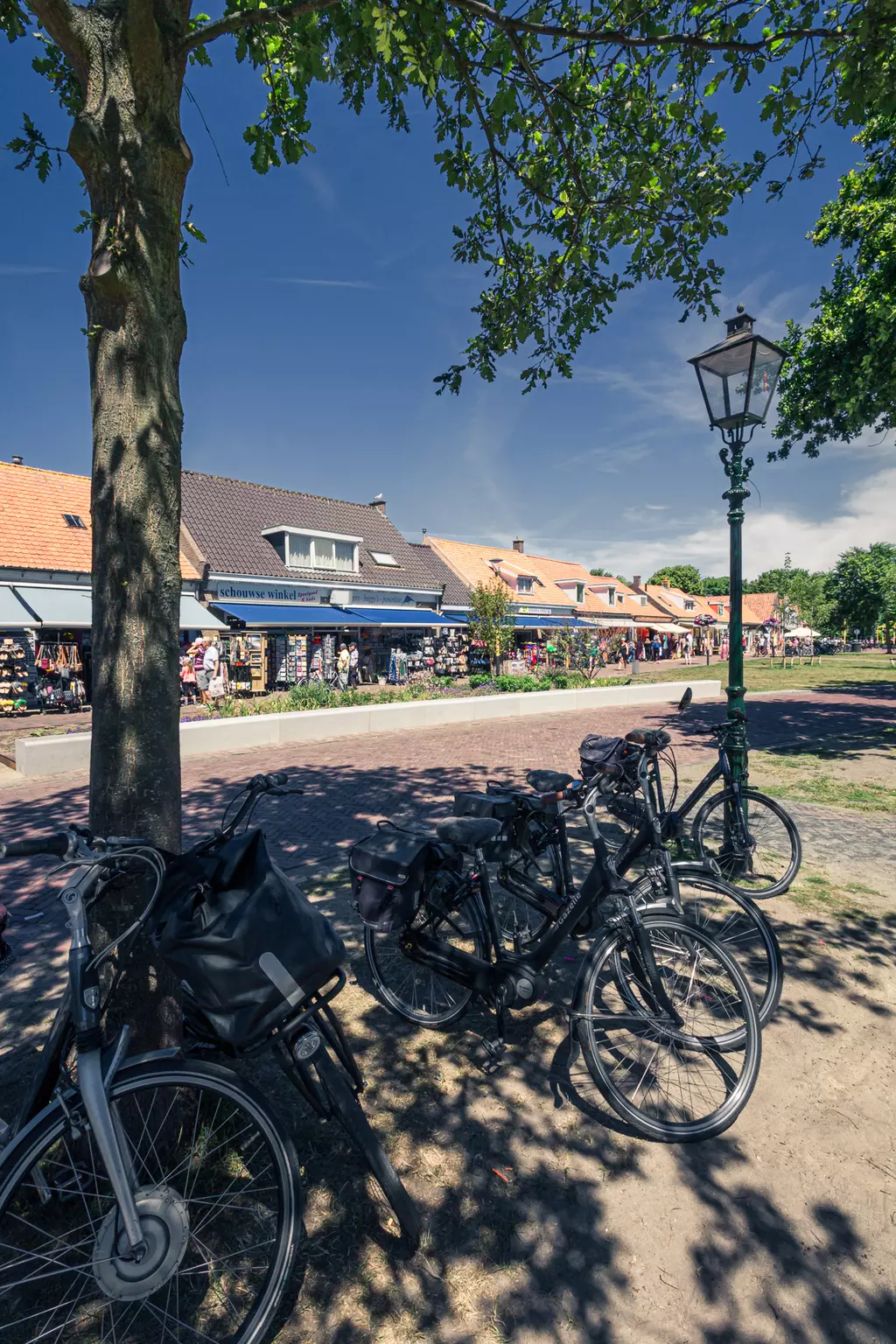 Bicycles are parked under a tree. In the background, shops display colorful items alongside a paved path.