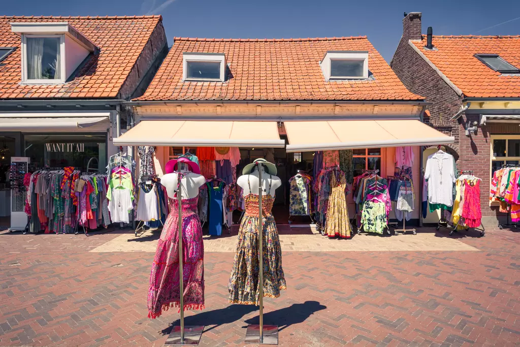 Two mannequins display colorful dresses in front of a store with sunny façades and a variety of clothing in the background.