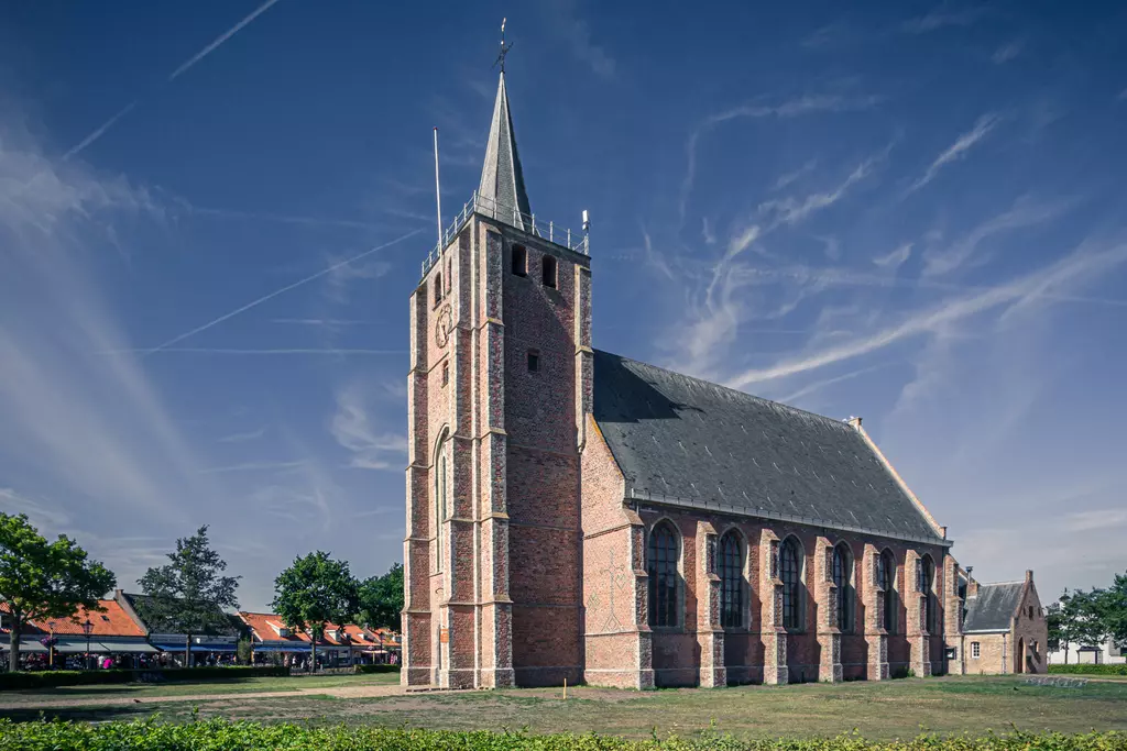 A brick building with a tall, pointed tower and large windows, surrounded by trees and grass.