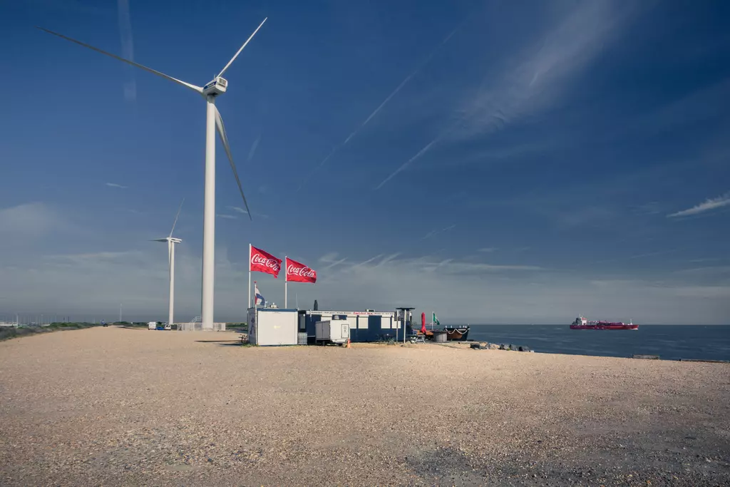 A gravel area with wind turbines in the background, two red flags fluttering, and a ship passing on the water.