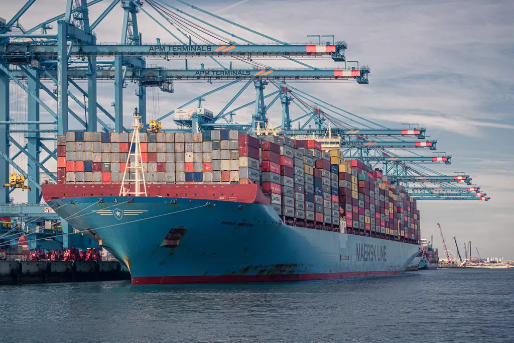 A large cargo ship rests in the harbor, loaded with colorful containers and surrounded by crane systems.