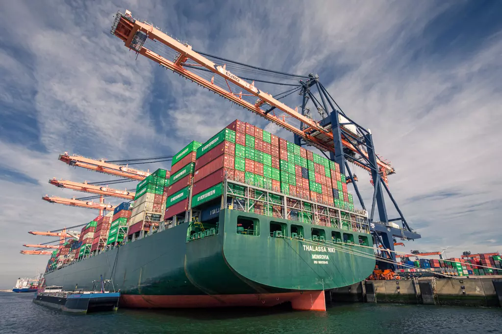 A large cargo ship with colorful containers is being unloaded by cranes at the port, with water in the foreground and a clear sky.