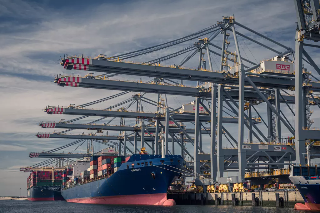 Container ships are docked at a busy port with large cranes looming in the background under a blue sky.