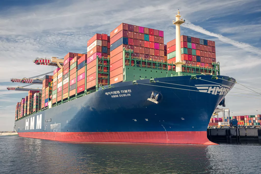 A large container ship loaded with colorful containers in the harbor. Water reflects the ship and the clear sky.