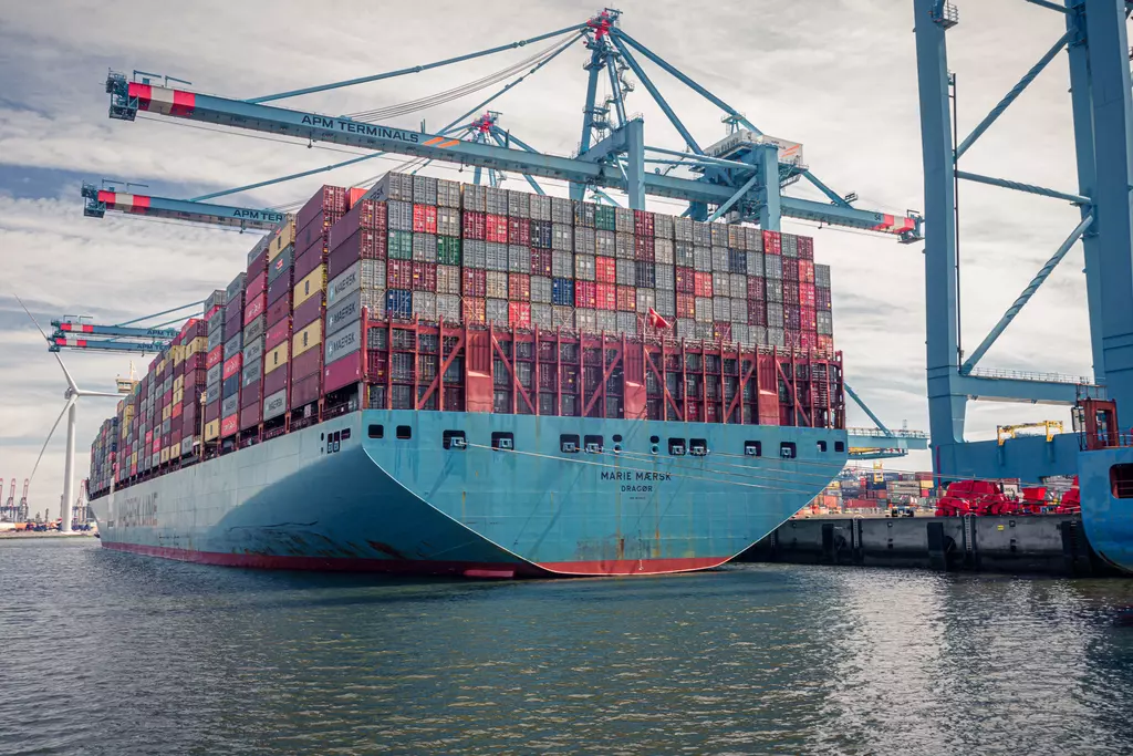 A large cargo ship with colorful containers in the harbor, surrounded by cranes and water.