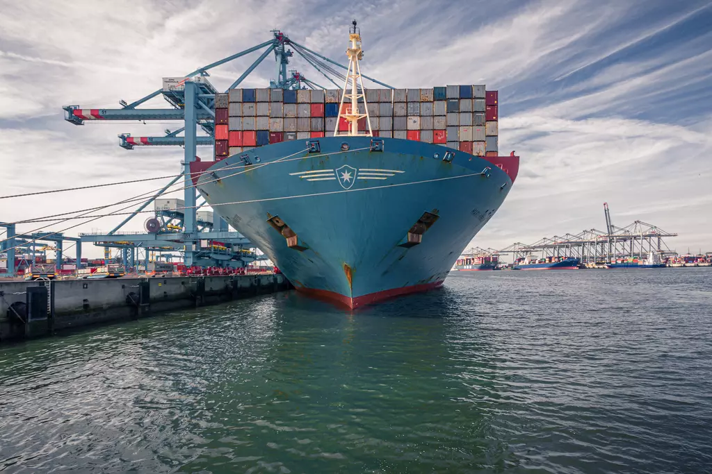 A large cargo ship is docked in the water, loaded with colorful containers. Crane structures are visible in the background.