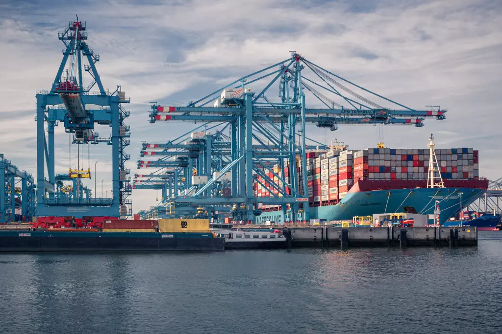 A port featuring large cranes, container ships, and water in the foreground, with many colorful containers visible.