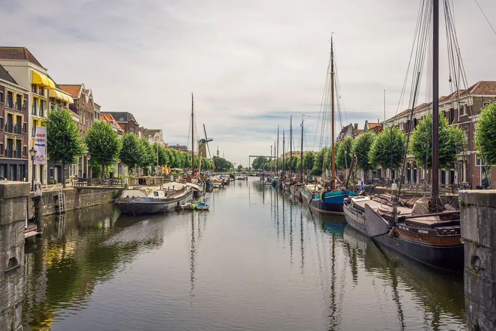 A calm canal with boats, lined by houses and trees on both sides, reflecting the clouds on the water.