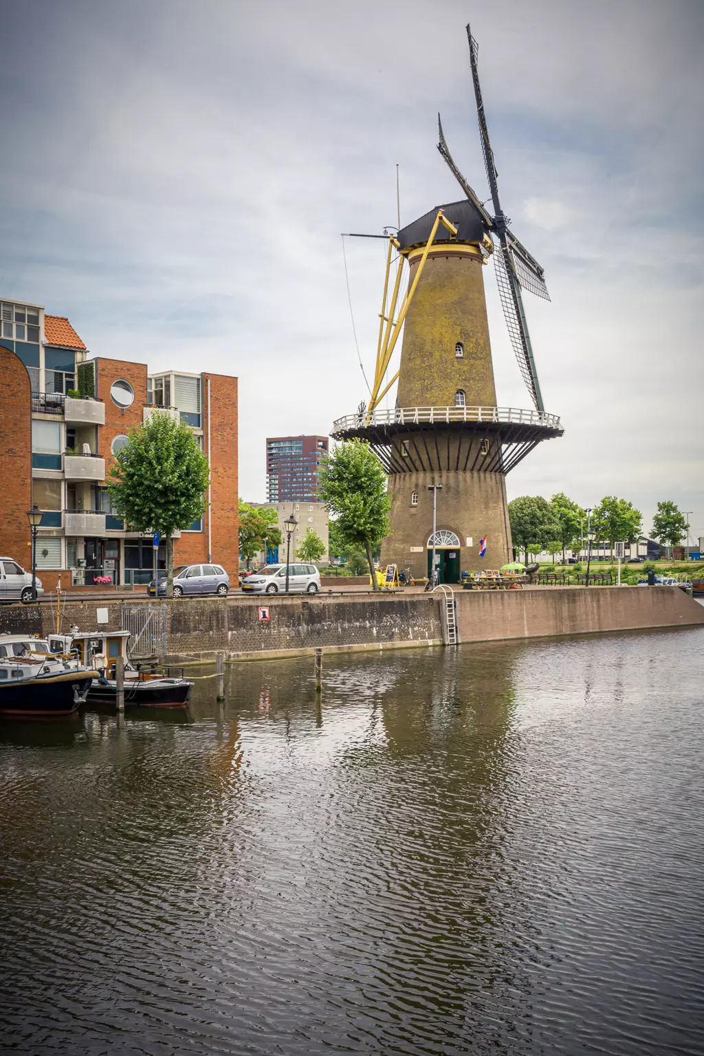 A windmill with large blades stands by the edge of a calm body of water, surrounded by modern buildings and trees.