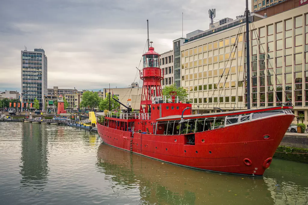 A red ship with a tall tower rests in a calm waterway, surrounded by modern buildings and urban life.