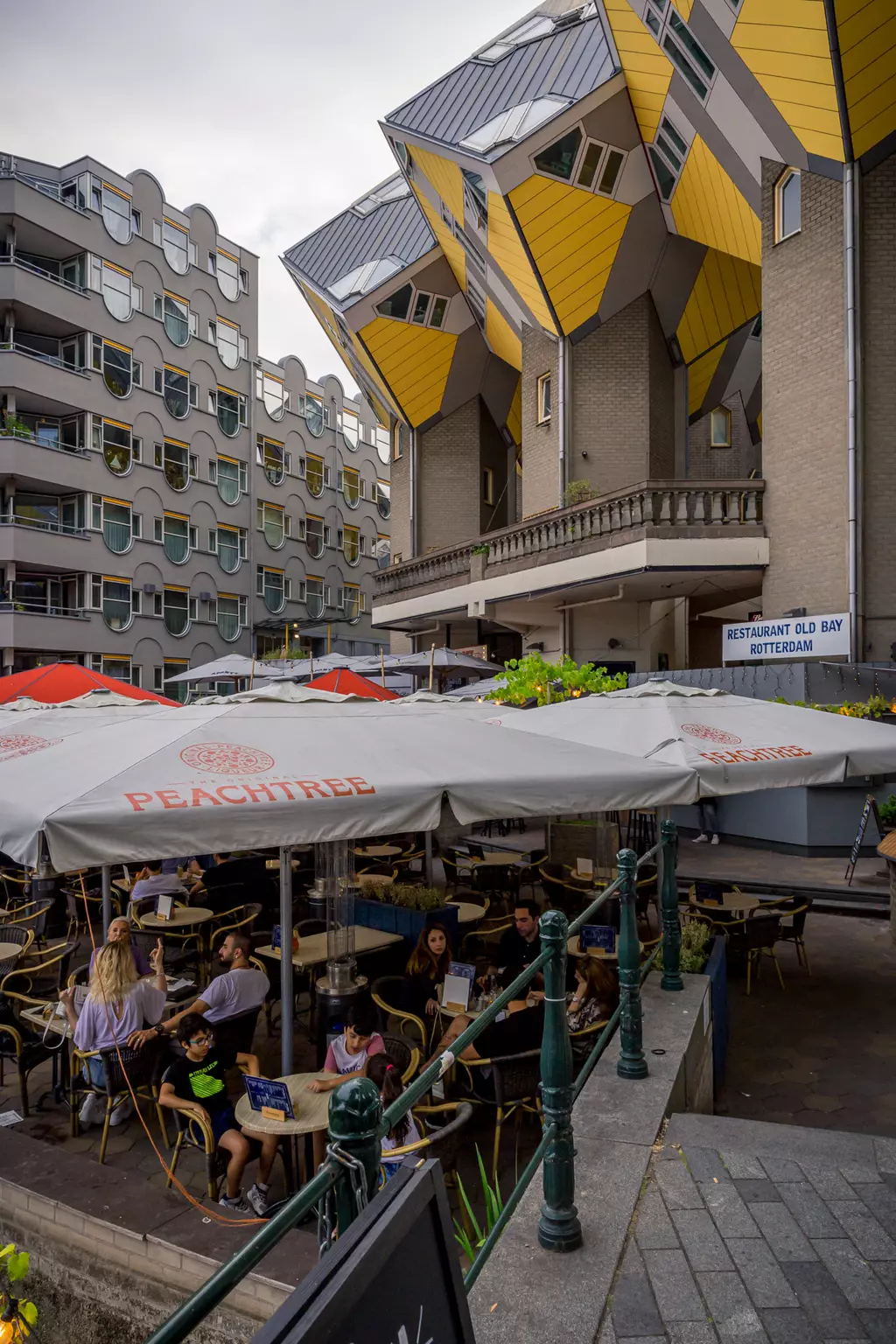 A terrace with tables and chairs, where people are dining and sitting. Bold modern buildings in the background.