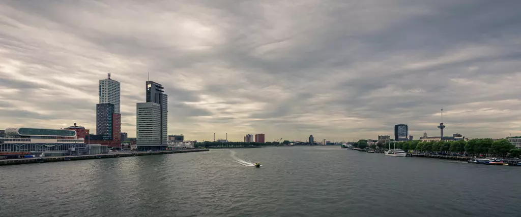 View of a cityscape with modern buildings along the river, a small boat crosses the water, and the sky is cloudy.