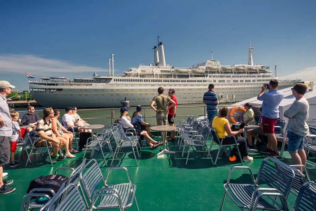 A group of people sits on a tour boat, with a large ship in the background under a clear blue sky.