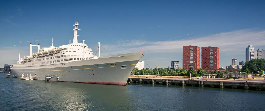 A large ship is docked at a harbor. In the background, modern buildings and trees are visible. The sky is clear and blue.