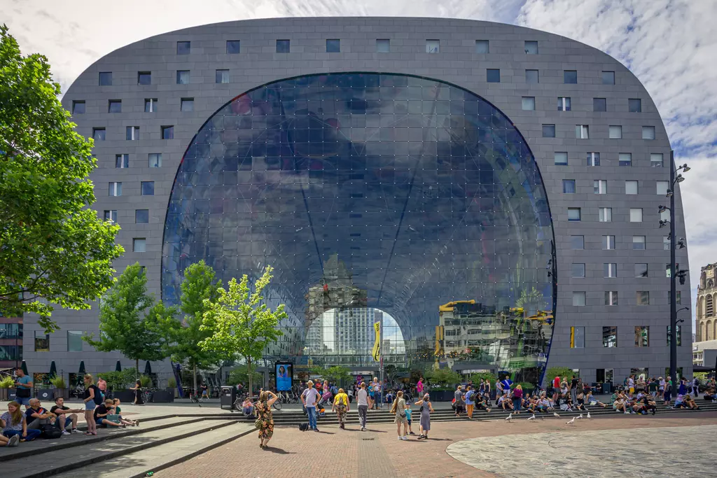 A modern, arch-shaped building with a glass facade, surrounded by trees and people gathering in the plaza.