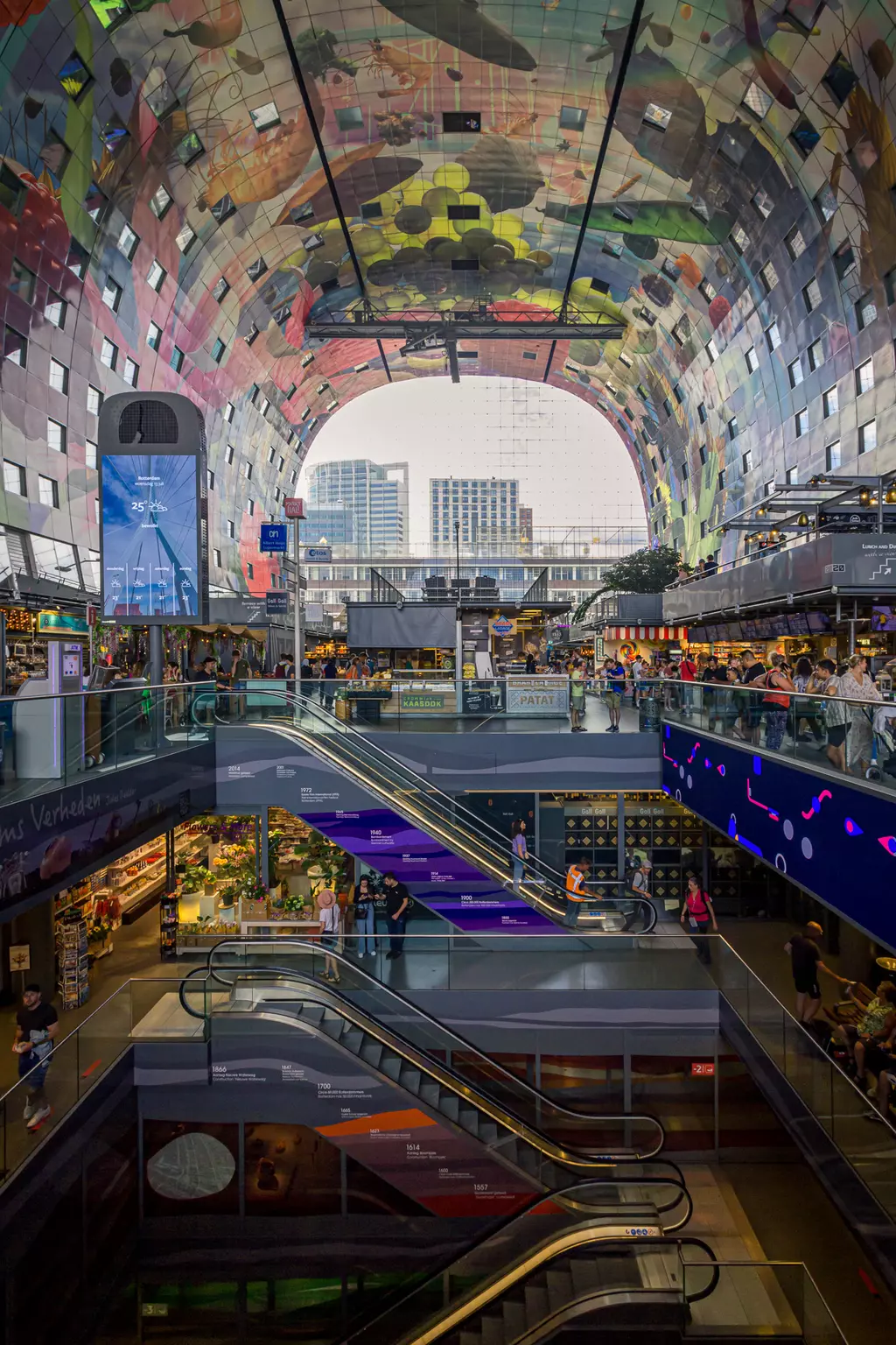 A colorful vaulted ceiling over a busy market hall. People are moving on escalators between shops and stalls.