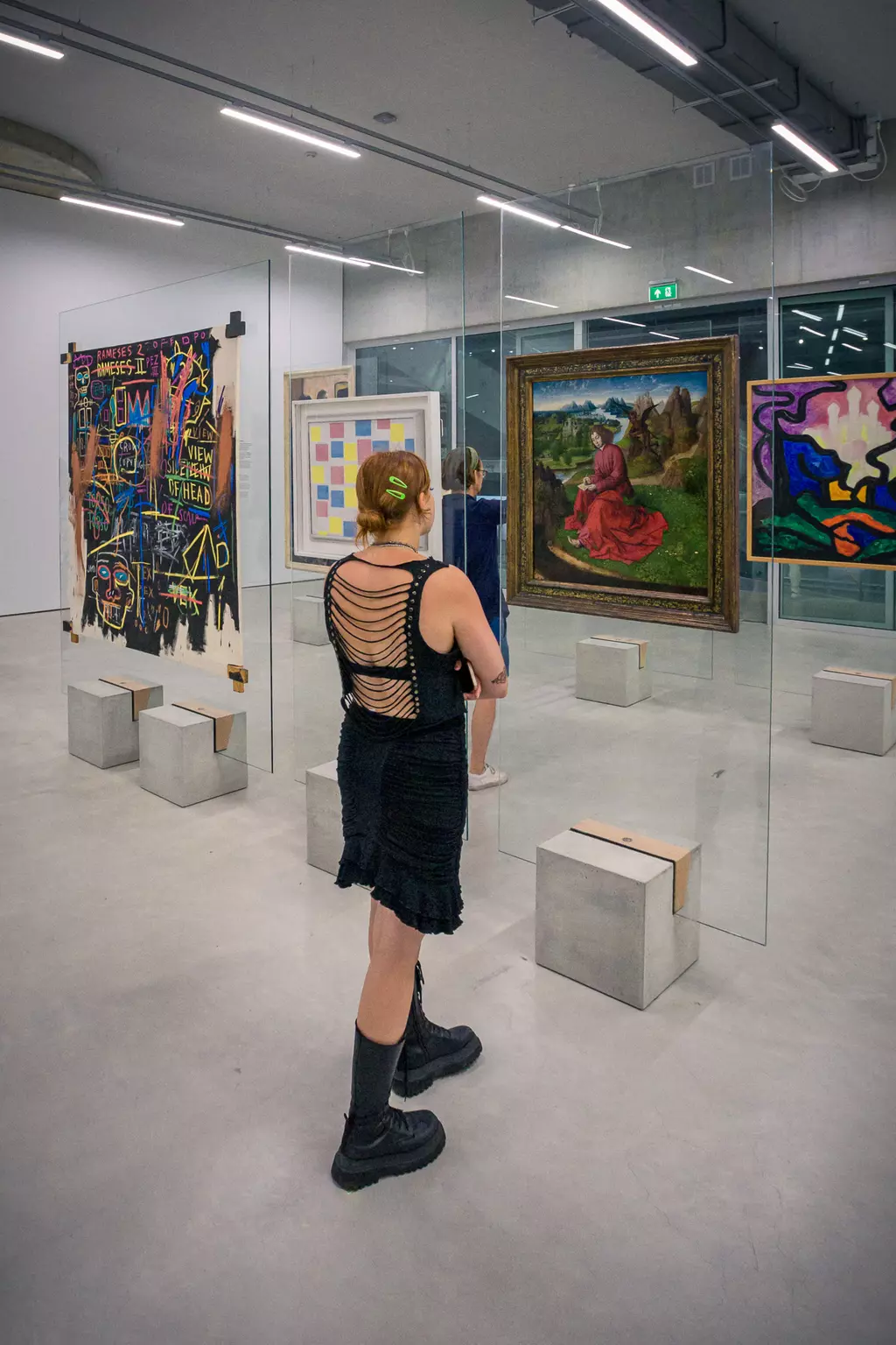 A person in a black dress and boots looks at artworks in a gallery. Several paintings are displayed on glass partitions.