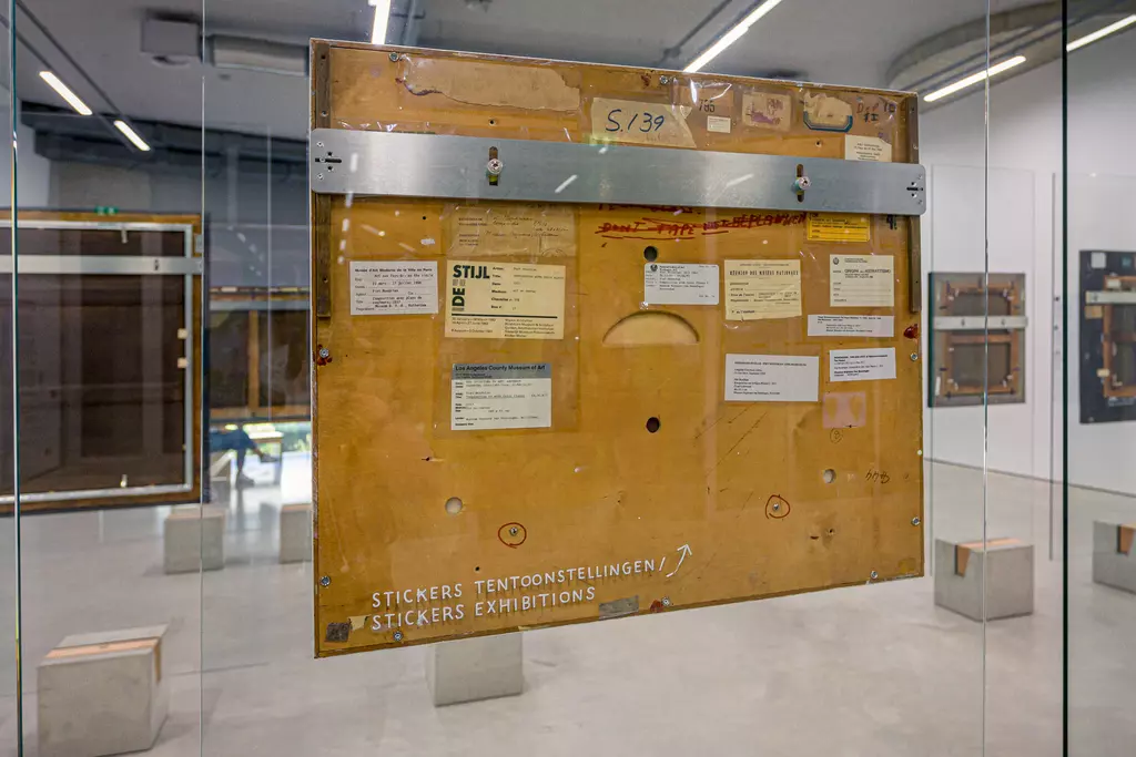 A wooden exhibition board with stickers and labels hangs behind glass in a modern gallery space.