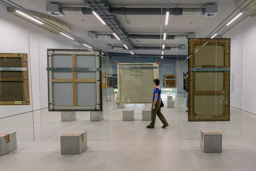 Two large framed artworks hang in front of transparent walls. A person walks between the exhibits, surrounded by neutral decor.