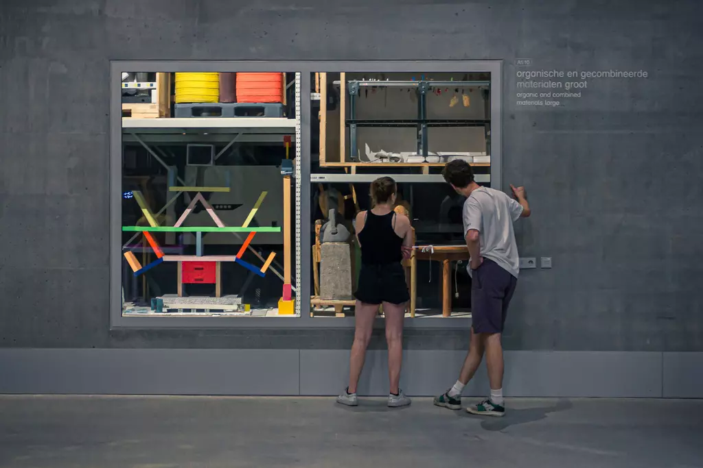 Two people are looking at a large wall display filled with colorful, organically arranged objects and materials in a modern space.