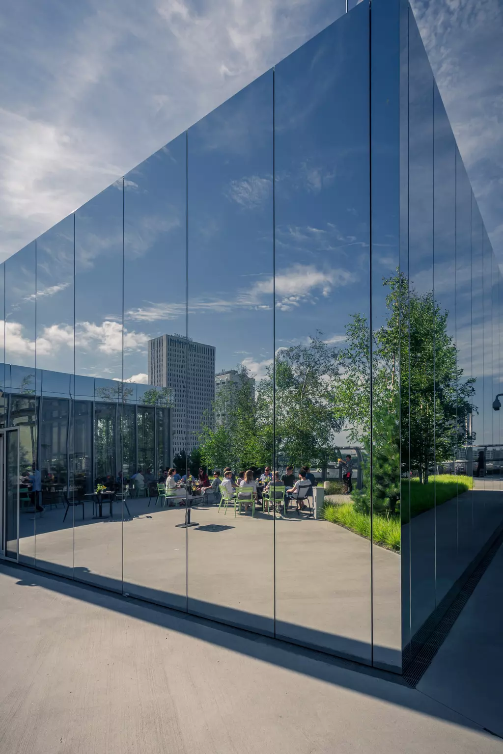 Reflective glass walls surround a terrace with people, plants, and benches. The sky is partly cloudy.