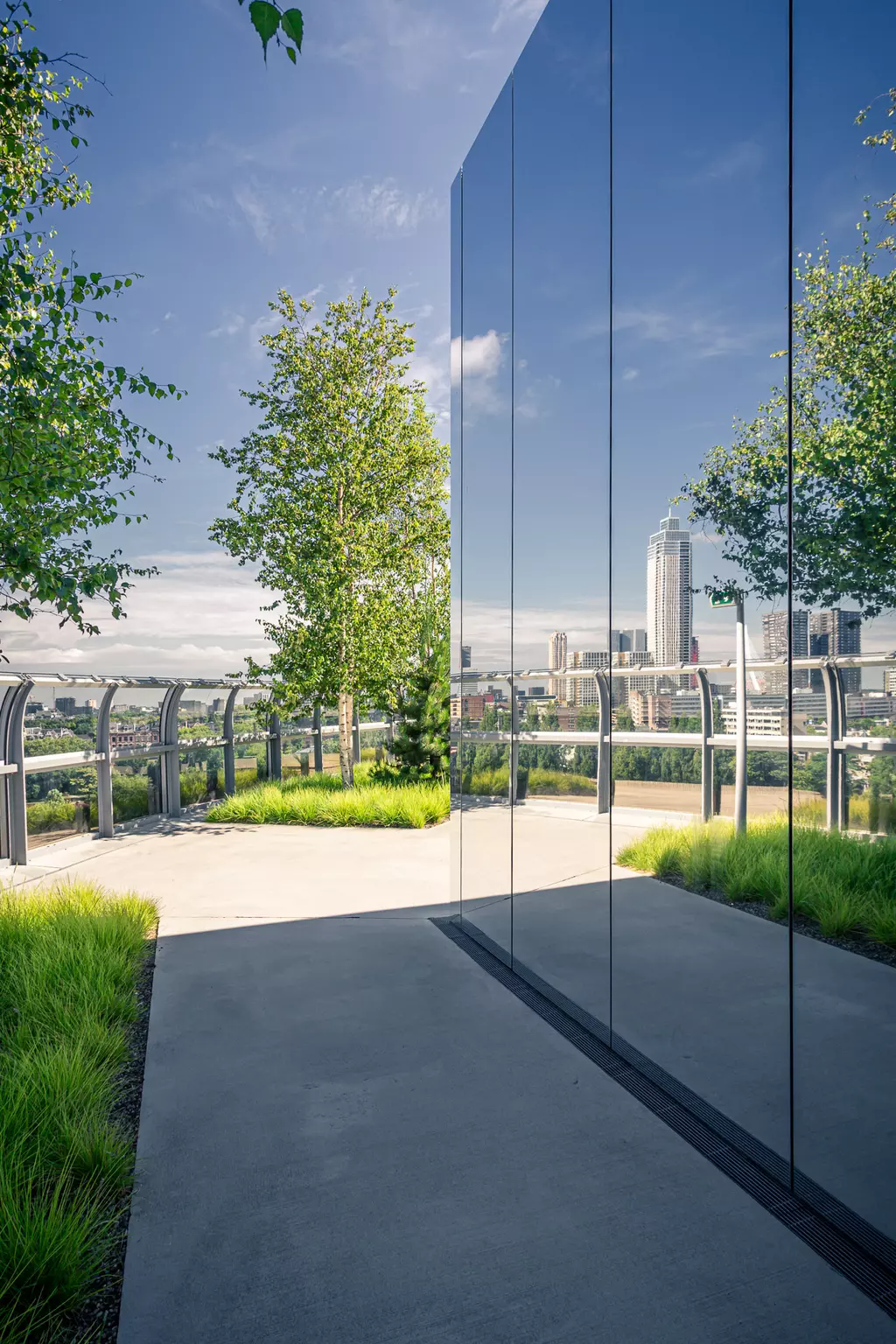 A paved path leads between tall mirrored walls and trees, with a city skyline in the background.