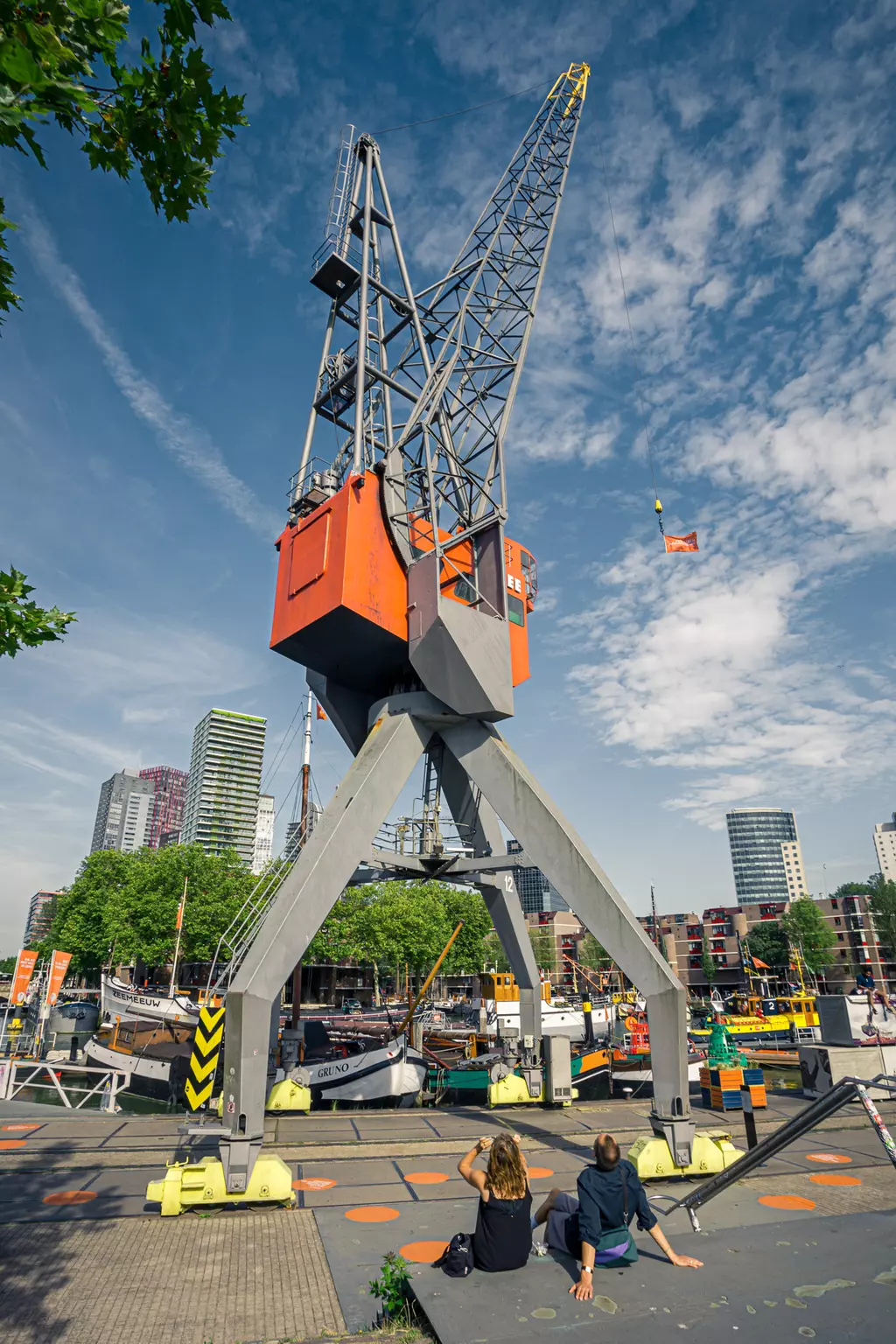 Two people sit at the waterfront as a large crane looms overhead, lifting a flag with modern buildings in the background.