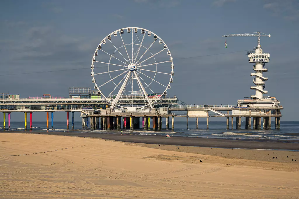 A large Ferris wheel stands beside a pier extending into the sea, with a sandy beach spread out in the foreground.