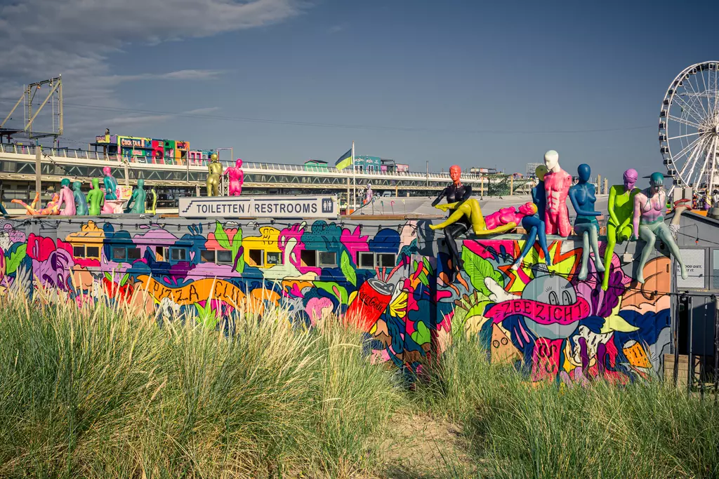 Colorful murals with figures adorn a building, surrounded by tall grass and a Ferris wheel in the background.