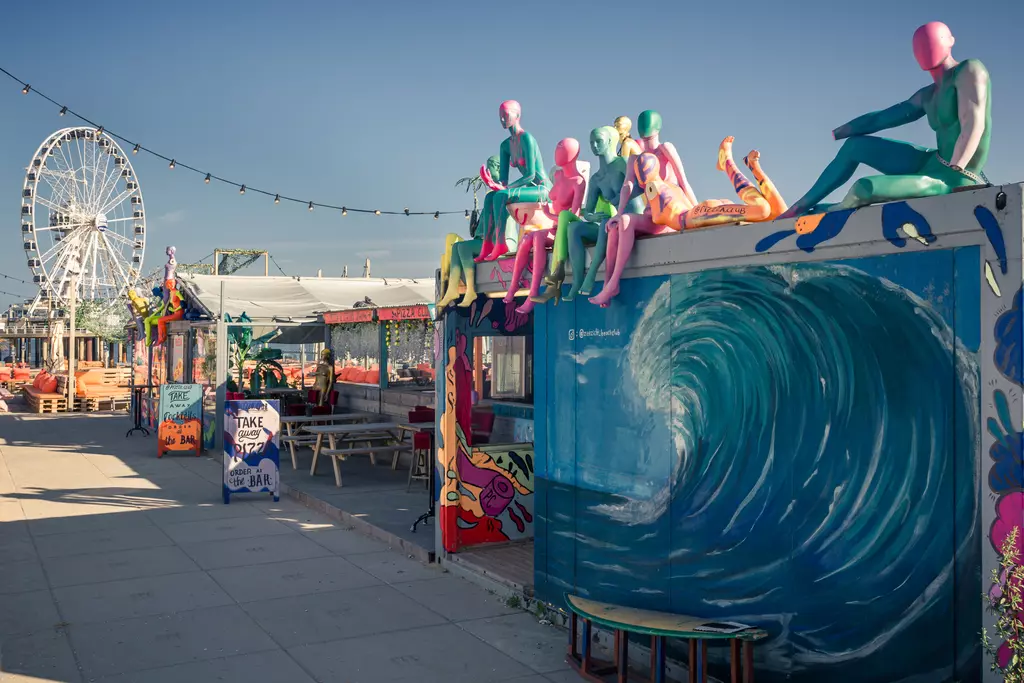 Colorful sculptures sit atop containers; a large wave mural and a ferris wheel are in the background under a clear sky.