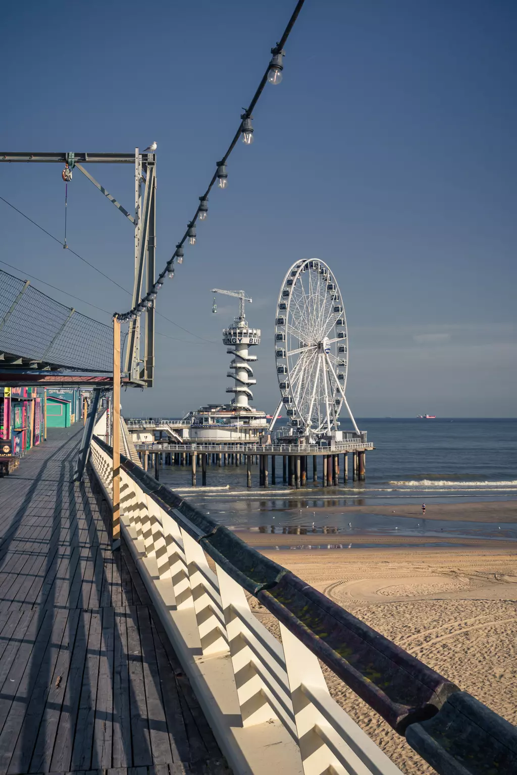 A wooden walkway leads along the sea to a pier with a large ferris wheel and a tower in the background.