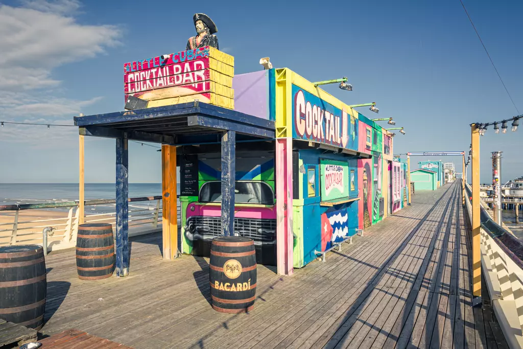 Colorful cocktail bar with large signs, wooden planks, and barrels on a pier, with a sea view in the background.