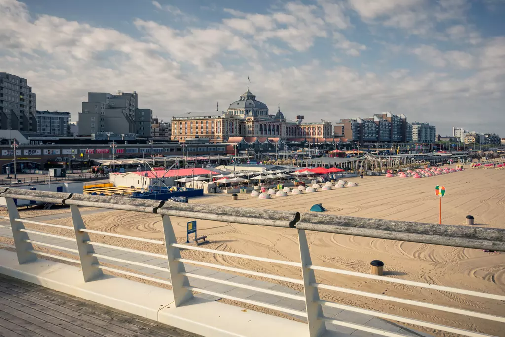 Sandy beach with deck chairs and umbrellas, flat buildings and clouds in the background.