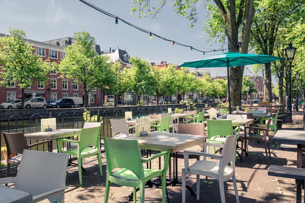 Colorful tables and chairs are set in a café area surrounded by trees, with a canal in the background.