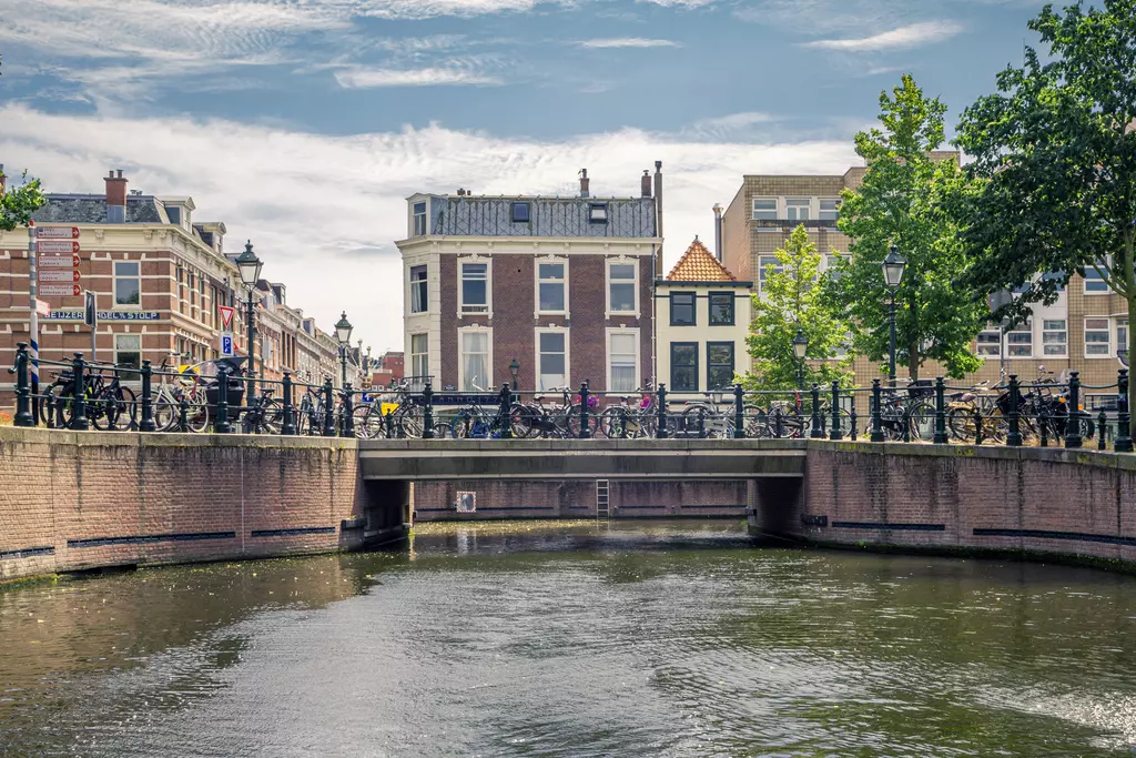 A calm water surface surrounded by historic buildings and many bicycles, with trees and a bright sky above.