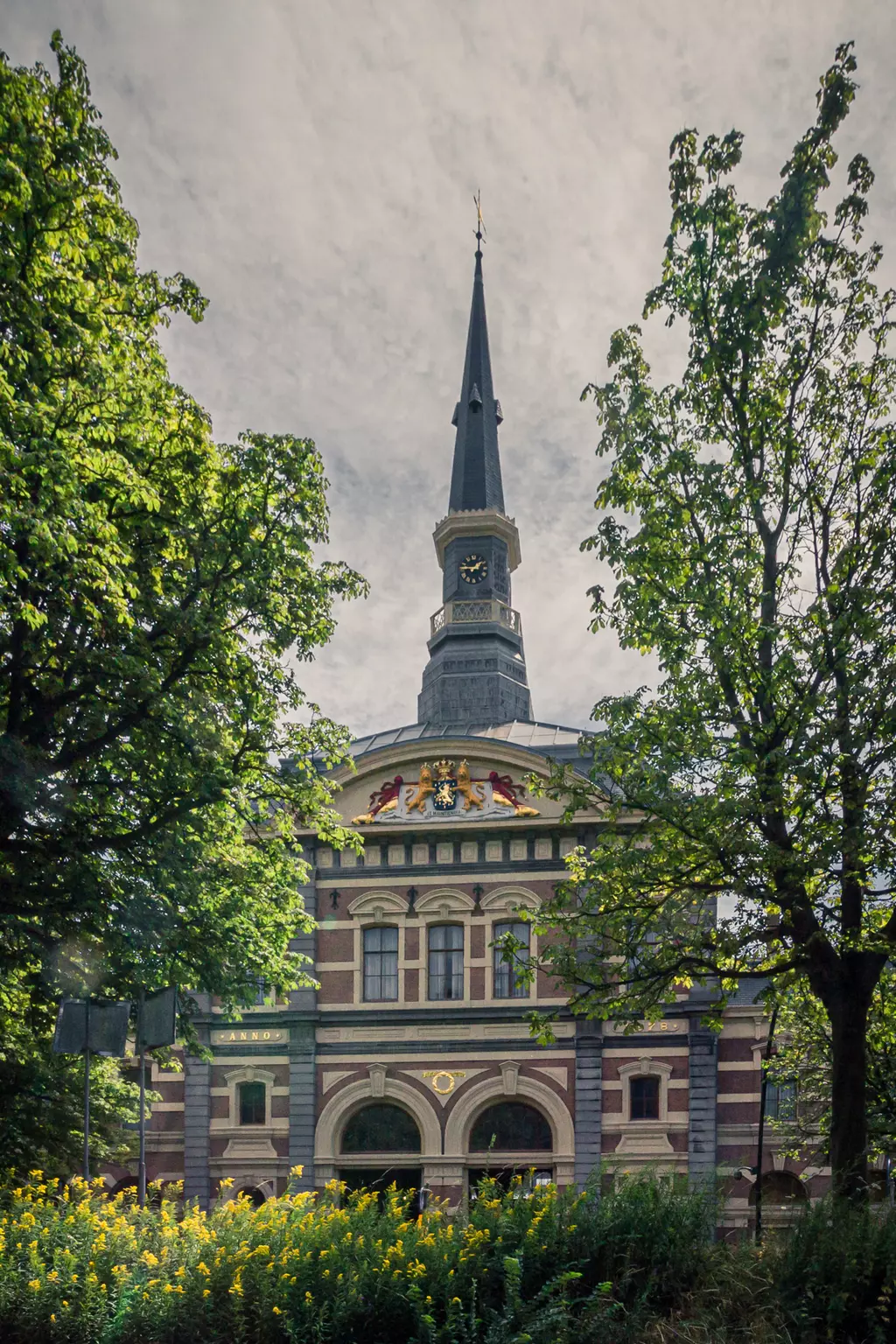 A view of a historic building with a tall pointed tower and colorful ornaments, surrounded by trees and yellow flowers.