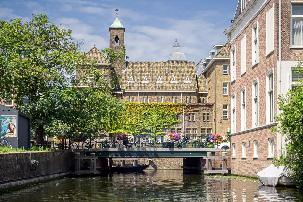 A canal with a small bridge, surrounded by historic buildings, trees, and blooming plants, with boats floating on the water.