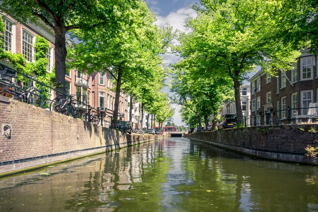 Green trees line a serene canal, with houses featuring windows and bicycles along the banks. The water reflects the scenery.