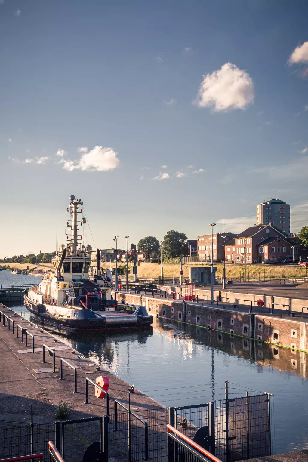 A tugboat rests in the harbor next to a calm waterway, surrounded by buildings and gentle hills under a blue sky.