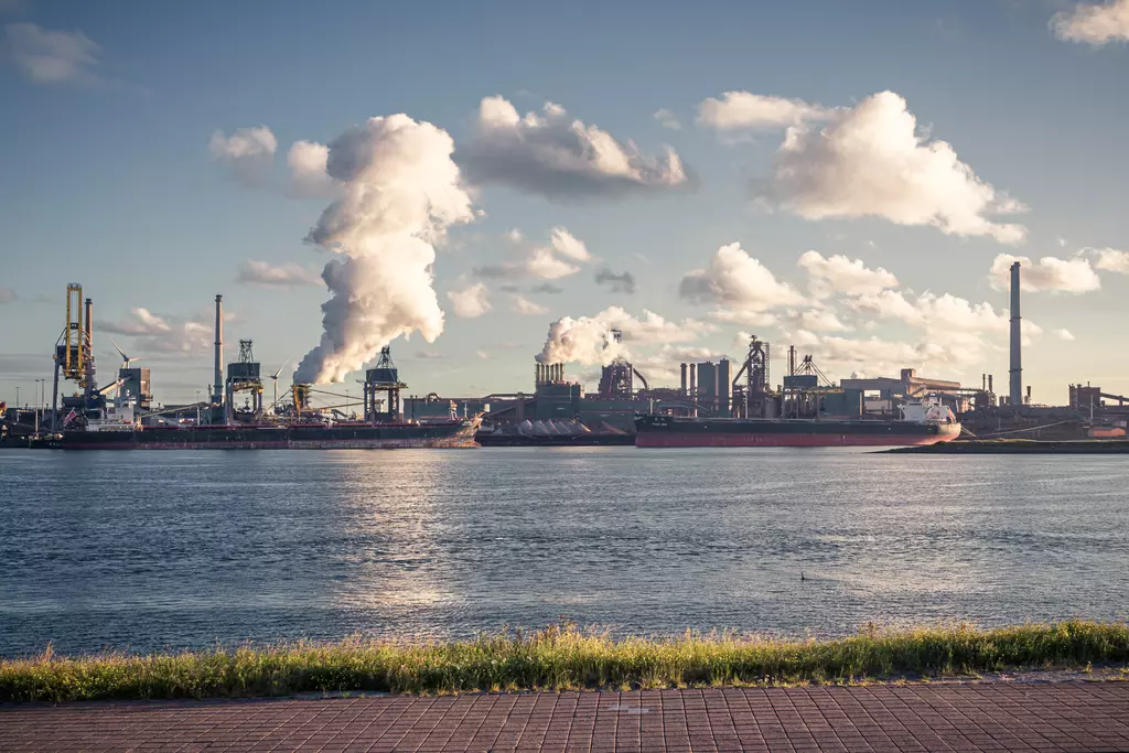 Large ships in the harbor, steam rising, factories in the background, calm water with a green grass line in the foreground.