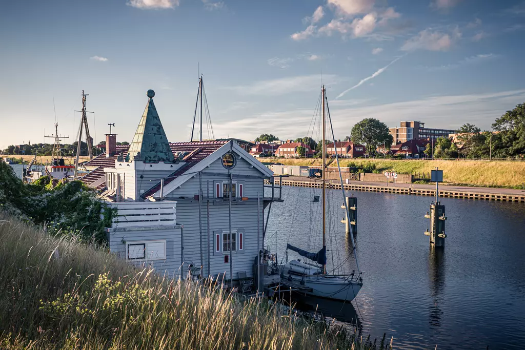 A white building with a green roof stands by a calm waterway, surrounded by boats and lush vegetation.