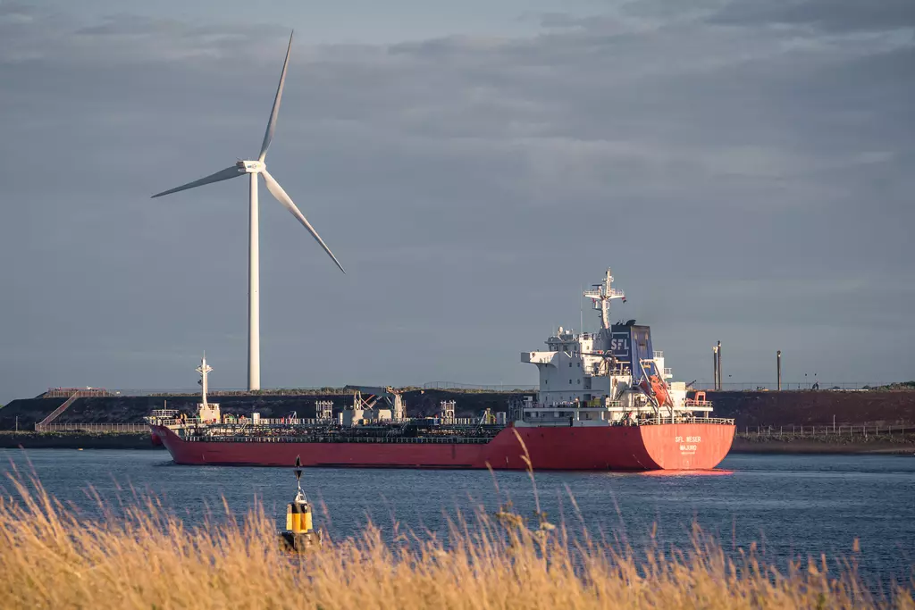 A large cargo ship sails in the water, passing by a wind turbine and lush riverbanks.