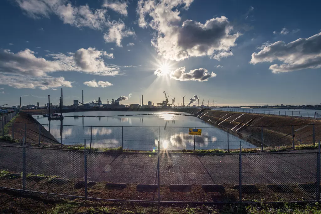 A water basin reflects the sun and clouds, surrounded by a fence and industrial structures in the background.