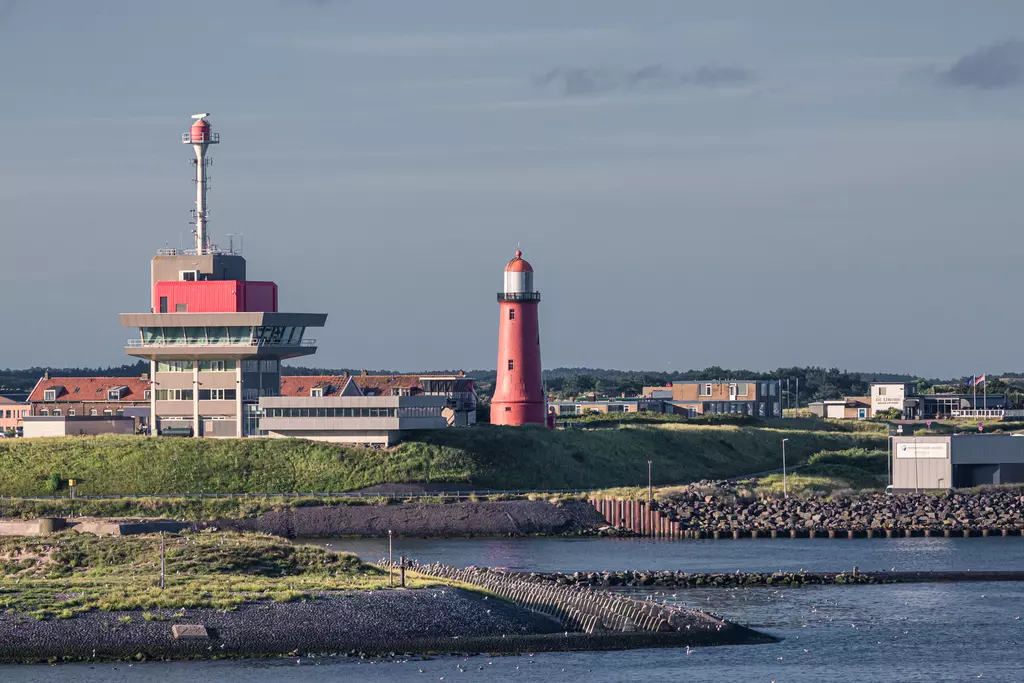 A red lighthouse and a modern building stand at a harbor entrance, surrounded by water and grassland.