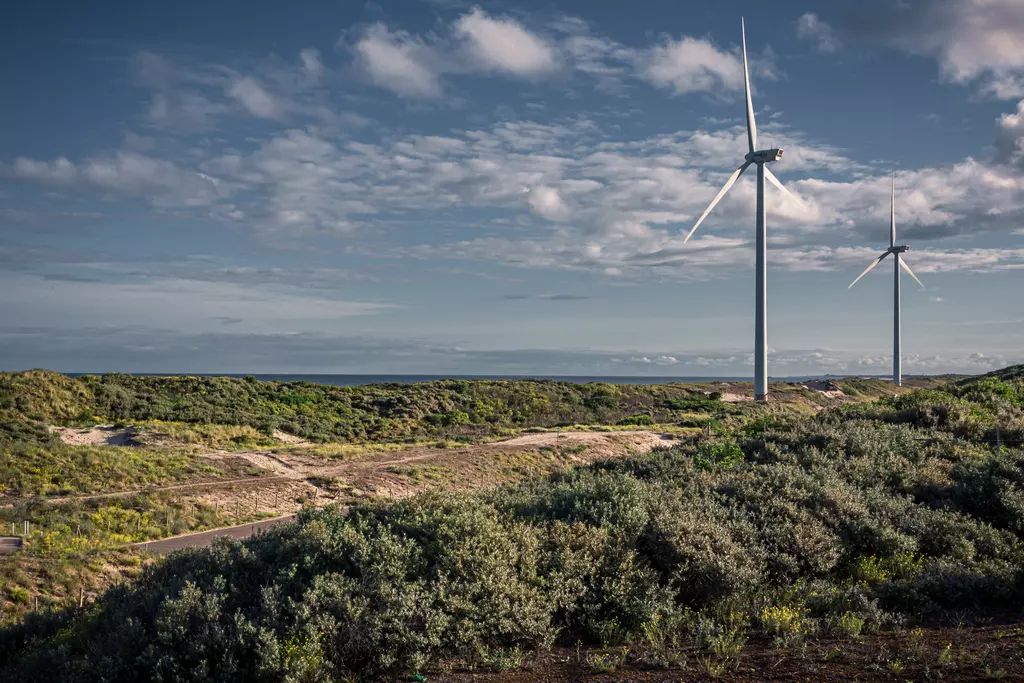 Two wind turbines tower over a hilly landscape filled with green shrubs and a clear sky in the background.