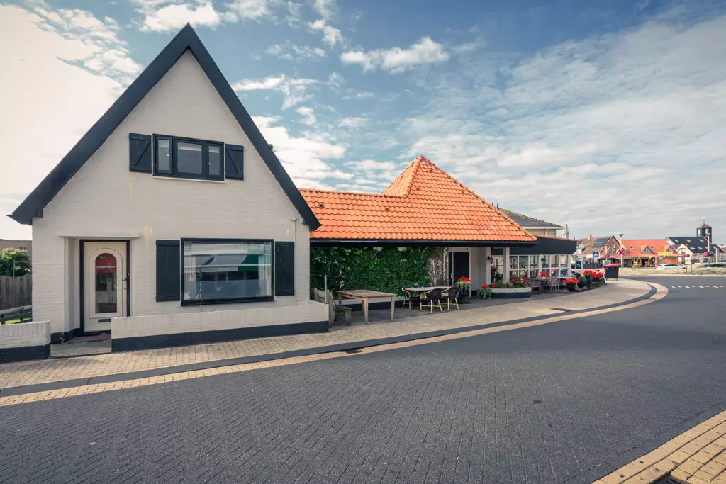 Haus in Callantsoog: Ein weißes Gebäude mit einem schwarzen Dach und roten Ziegeldächern, umgeben von blühenden Pflanzen und einem glatten Straßenverlauf.