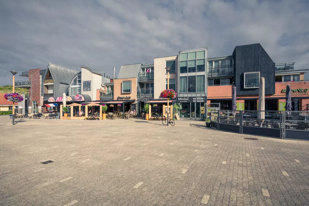 Einkaufsstraße in Callantsoog: Moderne Gebäude mit Cafés und Blumen, gepflasterter Platz mit Fahrrad und wenigen Passanten.