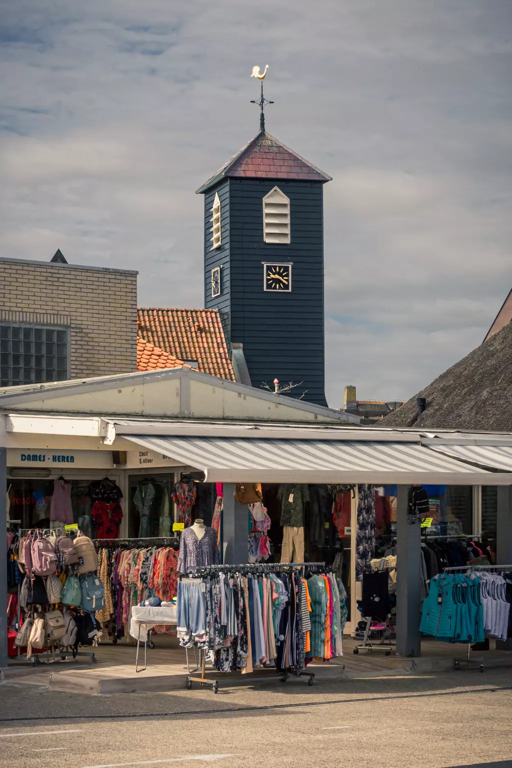 Markt in Callantsoog: Ein blauer Uhrturm mit Dachspitze über einem Bekleidungsgeschäft mit Kleidung auf Ständern.