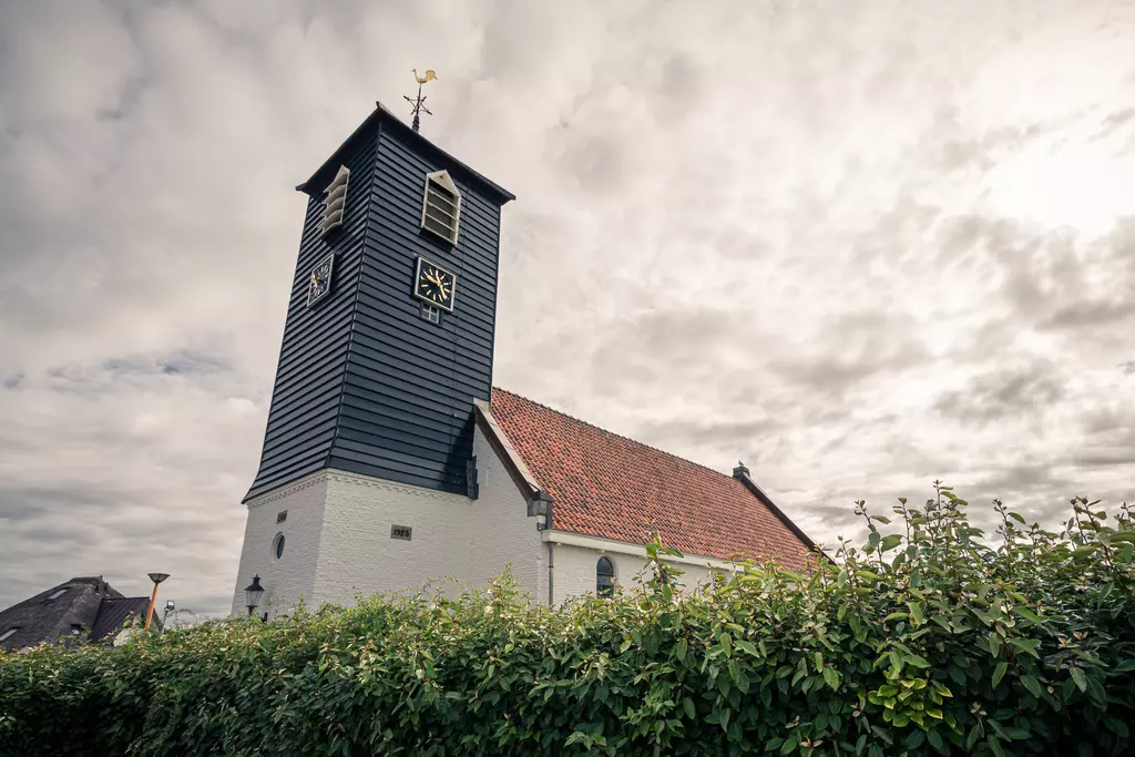 Kirche in Callantsoog: Ein schiefer Turm mit Uhr, umgeben von grünen Sträuchern, im Hintergrund ein bewölkter Himmel.