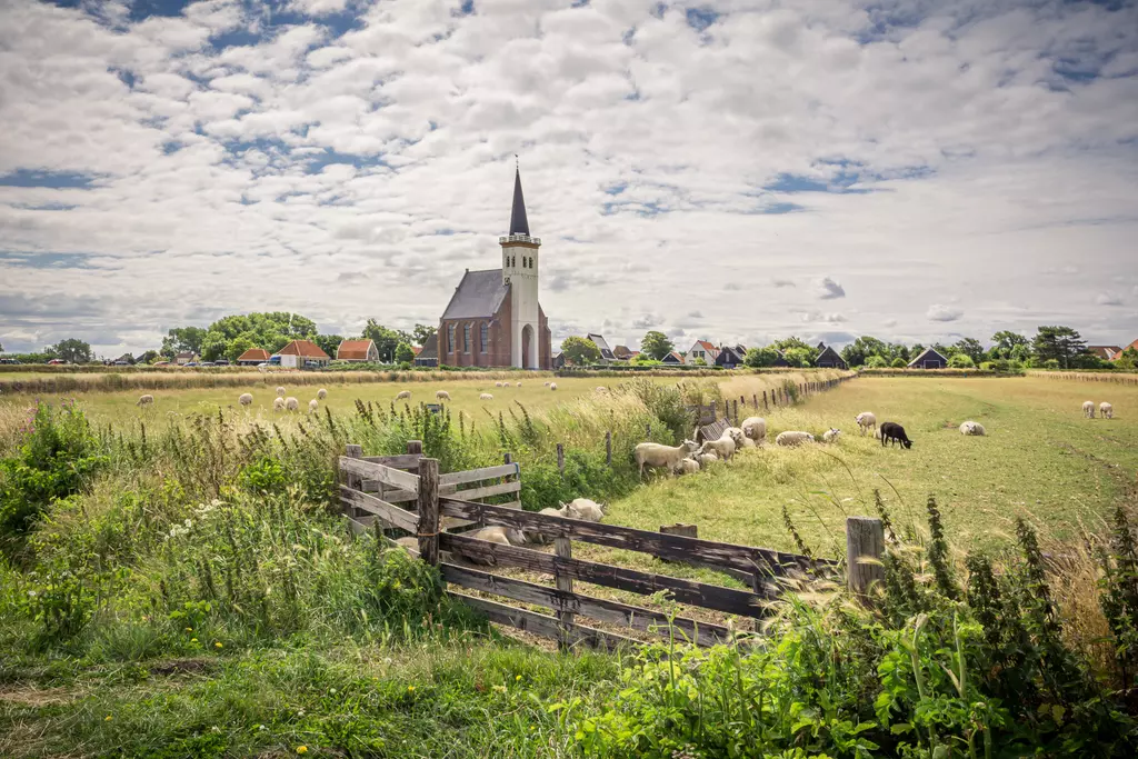 Eine Kirche mit spitzem Turm neben üppigen Wiesen, auf denen Schafe grasen, unter einem bewölkten Himmel.